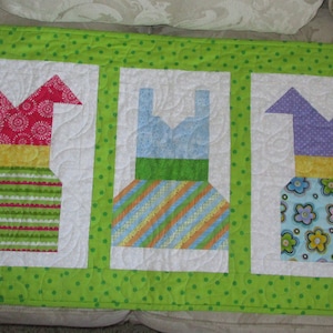 May include: A quilted wall hanging with five colorful fabric dresses sewn on it. The dresses are in shades of red, blue, green, purple, and pink. Each dress has a different pattern and is framed with a white border.