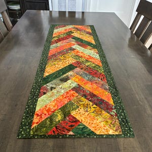 May include: A rectangular table runner featuring a herringbone pattern made of quilted fabric strips in shades of orange, red, yellow, and green. The runner is bordered by a dark green fabric with a subtle, lighter green speckled print throughout.