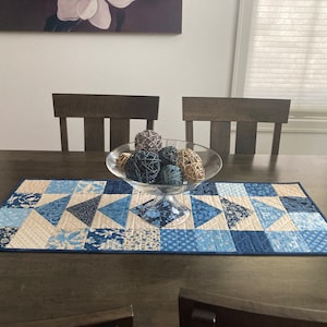 May include: A blue and white patchwork table runner with a blue border sits on a dark wood table. A glass bowl filled with decorative balls sits in the center of the runner.