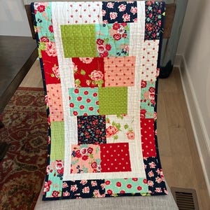 May include: A colorful patchwork quilt with a variety of floral and patterned fabrics. The quilt features red, green, pink, and navy blue squares, with a white border and backing. The quilt is approximately 40 inches long.