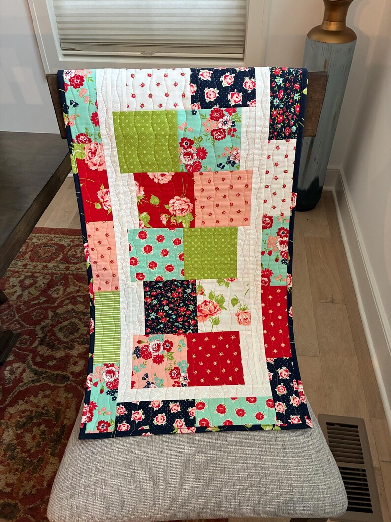 May include: A colorful patchwork quilt with a variety of floral and patterned fabrics. The quilt features red, green, pink, and navy blue squares, with a white border and backing. The quilt is approximately 40 inches long.