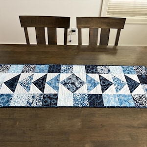 May include: A rectangular quilted table runner featuring a patchwork design in various shades of blue and white. The pattern consists of geometric shapes, including triangles and squares, with floral and abstract motifs printed on the fabric.