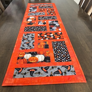 May include: A rectangular quilted table runner featuring a vibrant orange border and a patchwork design of Halloween-themed fabric blocks. The patterns include black witch hats, bats, spiderwebs, and various orange and white pumpkins on black backgrounds.