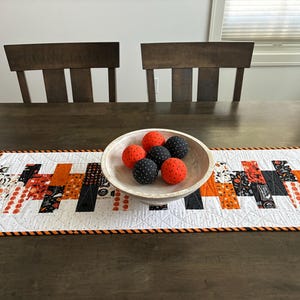 May include: A Halloween-themed table runner with a white background and orange, black, and patterned fabric blocks. A wooden bowl holds six decorative balls, three orange and three black with white polka dots. The runner is on a dark wooden table.