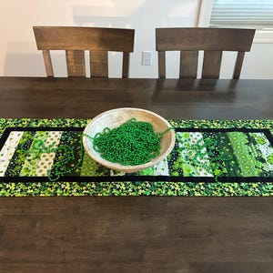 May include: A St. Patrick's Day themed table runner with a patchwork design in shades of green and white, featuring shamrocks and floral patterns. A wooden bowl filled with green beads and a strand of green beads are on the runner.