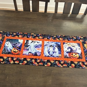 May include: A rectangular fabric table runner featuring a Halloween theme with four central panels depicting ghosts, jack-o'-lanterns, and haunted houses. The border is orange with black polka dots, surrounded by a black fabric with pumpkin prints.