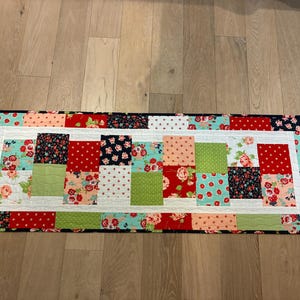 May include: A rectangular quilted table runner with a patchwork design. The runner features squares of red, green, pink, and navy fabrics with floral and polka dot patterns. A white border frames the colorful center.