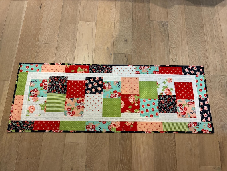 May include: A rectangular quilted table runner with a patchwork design. The runner features squares of red, green, pink, and navy fabrics with floral and polka dot patterns. A white border frames the colorful center.