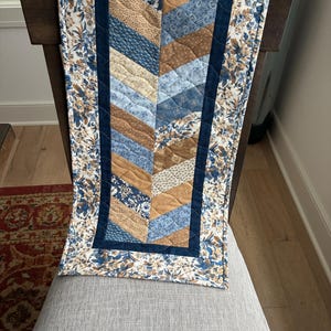 May include: A quilted table runner with a chevron pattern in shades of blue, tan, and cream. The runner is bordered with a floral print and a dark blue trim. It is draped over the back of a wooden chair.