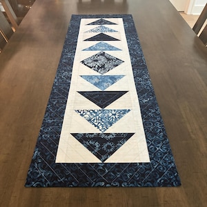 May include: A rectangular quilted table runner featuring a central row of alternating navy blue and white triangles and diamond shapes with batik-style patterns. The runner is framed by a dark blue border with a repeating swirl design and white stitching.