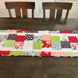 May include: A rectangular patchwork table runner with a variety of colorful floral and patterned fabrics. The runner features squares of red, pink, green, and navy blue fabrics, bordered by a navy blue edge. The runner is placed on a dark wooden table.