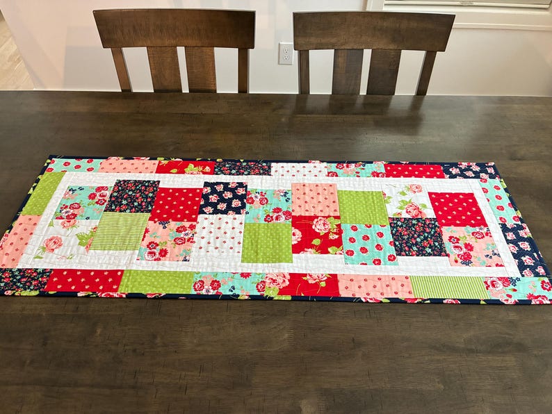 May include: A rectangular patchwork table runner with a variety of colorful floral and patterned fabrics. The runner features squares of red, pink, green, and navy blue fabrics, bordered by a navy blue edge. The runner is placed on a dark wooden table.