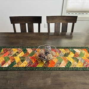 May include: A rectangular quilted table runner featuring a vibrant herringbone pattern in shades of orange, yellow, red, and green. The runner is bordered by a dark green fabric with a subtle pattern and holds a clear bowl with decorative spheres.
