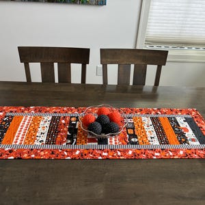 May include: A rectangular fabric table runner featuring a patchwork design with orange, black, and white Halloween-themed patterns, including pumpkins and ghosts. A clear glass bowl sits in the center, filled with orange and black fabric spheres.