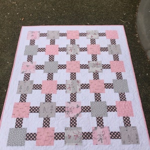 May include: A pink and gray patchwork quilt with a white background. The quilt is made up of squares of different fabrics, including polka dots, floral patterns, and solid colors. The quilt is edged with a pink border.