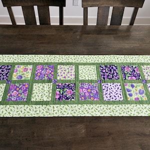 May include: A rectangular quilted table runner with a floral pattern. The runner features alternating squares of purple and light green floral fabric, framed by a green border with leaf designs. The runner is placed on a dark wooden table.