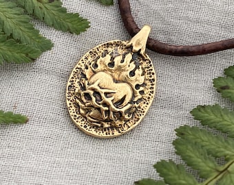 May include: A gold-coloured pendant necklace with an oval-shaped pendant featuring a detailed design of a skull and flames. The pendant hangs from a brown leather cord. Fern leaves surround the necklace.