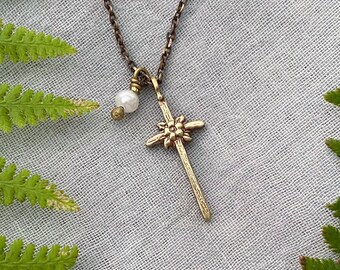 May include: A gold-toned pendant necklace with a cross design, floral detail, and a small pearl. The necklace is suspended from a fine chain. Green fern leaves and grey fabric form the background.