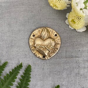Puede incluir: Un colgante de metal dorado con un corazón estilizado con una llama y espinas. El colgante está sobre un fondo de tela gris claro.