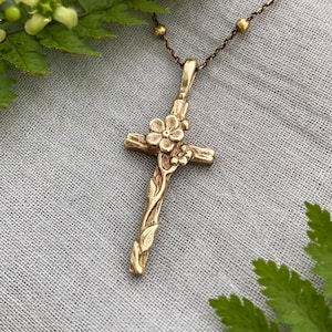 May include: A gold-coloured cross pendant necklace with floral detailing. The cross is adorned with delicate flowers and vines, and hangs from a delicate chain with small, round beads. The necklace is displayed on a grey surface, surrounded by green ferns and yellow flowers.
