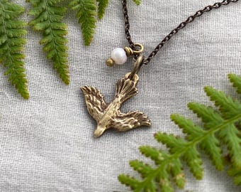 May include: A bronze-coloured pendant necklace with a stylized bird design. The bird pendant hangs from a delicate chain and is accented with a small, round, white pearl. The necklace is set against a backdrop of green fern leaves.