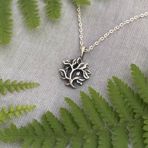 May include: A silver necklace with a tree pendant. The pendant is a detailed design of a tree with branches and leaves. The necklace is on a white background with green ferns.