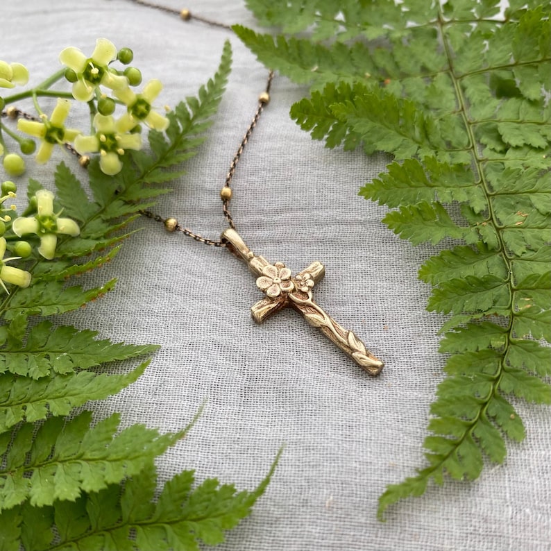 May include: A gold-coloured cross pendant necklace with floral and vine detailing. The cross is adorned with small flowers at the centre and along the arms. The necklace chain has small gold beads. The background includes green fern leaves and yellow flowers.