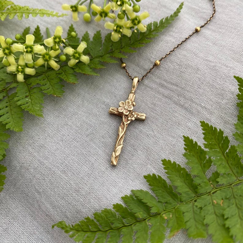 May include: A gold-coloured cross pendant necklace with floral detailing. The cross is adorned with delicate flowers and vines, and hangs from a delicate chain with small, round beads. The necklace is displayed on a grey surface, surrounded by green ferns and yellow flowers.