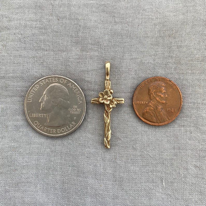 May include: A gold-coloured cross pendant with a floral design at the top and along the arms. The cross is displayed next to a quarter and a penny for size reference. The pendant is on a grey fabric surface.