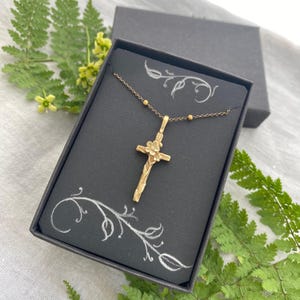 May include: A gold-coloured cross pendant necklace presented in a black gift box. The cross has a floral design and hangs from a fine chain. The box is decorated with white floral accents.