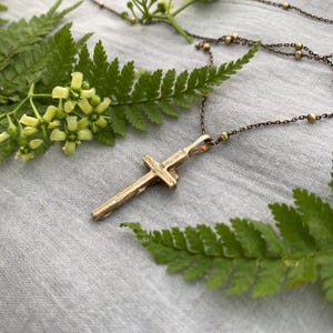 May include: A gold-coloured cross pendant necklace with a textured surface. The cross hangs from a delicate chain with small gold beads. The necklace is displayed on a grey fabric background, surrounded by green fern leaves and small yellow flowers.