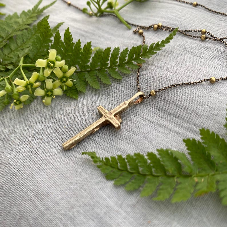 May include: A gold-coloured cross pendant necklace with a textured surface. The cross hangs from a delicate chain with small gold beads. The necklace is displayed on a grey fabric background, surrounded by green fern leaves and small yellow flowers.