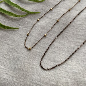 May include: Two delicate necklaces with a vintage bronze finish. One necklace features small, evenly spaced gold beads, while the other has a simple chain design. The necklaces rest on a light grey fabric, with a green fern leaf in the upper left corner.
