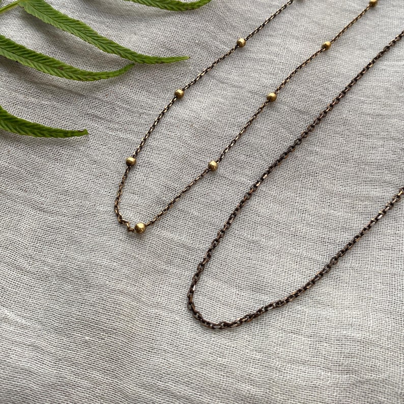May include: Two delicate necklaces with a vintage bronze finish. One necklace features small, evenly spaced gold beads, while the other has a simple chain design. The necklaces rest on a light grey fabric, with a green fern leaf in the upper left corner.