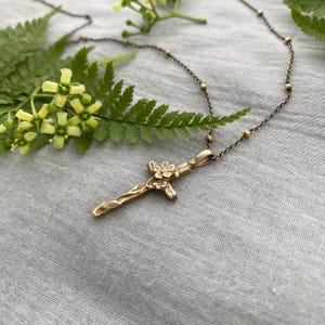 May include: A gold-toned pendant necklace with a cross design and floral detailing. The cross hangs from a fine chain with small, spherical beads. The necklace is displayed on a grey fabric surface, with a fern and flowers in the background.