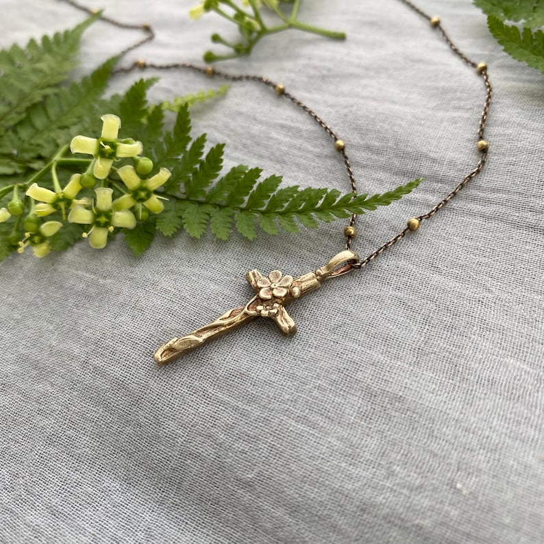 May include: A gold-toned pendant necklace with a cross design and floral detailing. The cross hangs from a fine chain with small, spherical beads. The necklace is displayed on a grey fabric surface, with a fern and flowers in the background.