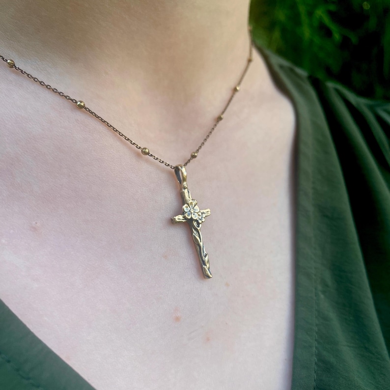 May include: A bronze-coloured pendant necklace. The pendant is a cross with a floral design. The cross has a textured, branch-like appearance. The necklace chain is delicate, with small, round beads.