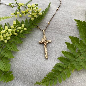 May include: A gold-coloured cross pendant necklace with floral detailing. The cross features a flower at the top and vines along the stem. The necklace chain has small gold beads. The image is set against a backdrop of green ferns and yellow flowers.