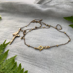 May include: A gold-coloured necklace featuring a cross pendant with a floral design. The chain is decorated with small gold beads and fastens with a circular clasp. The necklace is presented on a grey fabric background.
