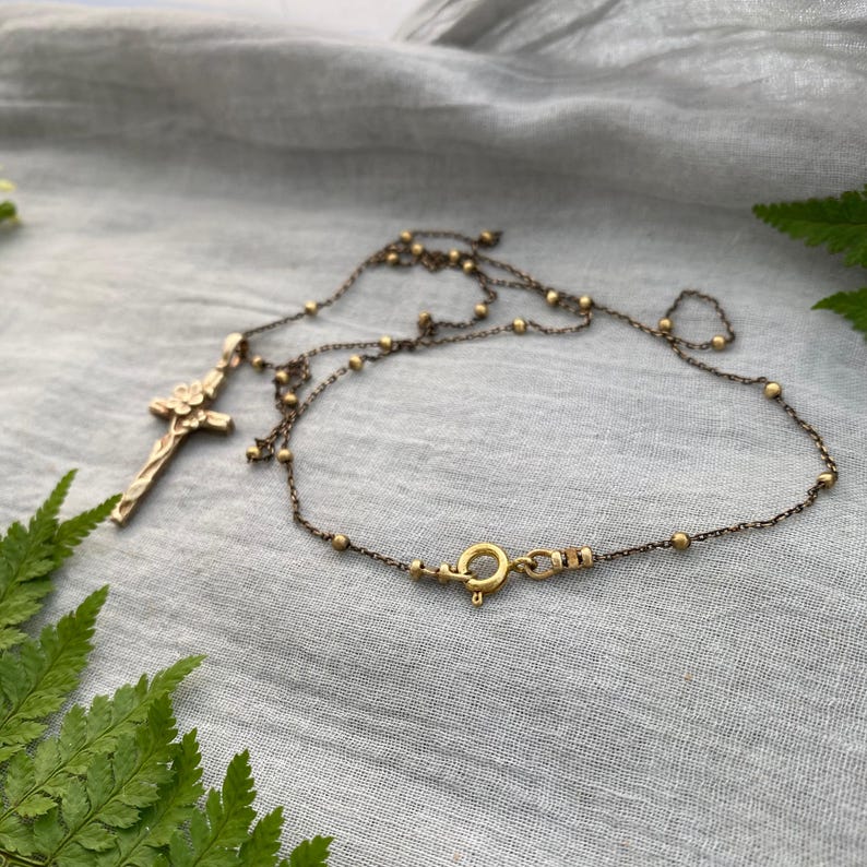 May include: A gold-coloured necklace featuring a cross pendant with a floral design. The chain is decorated with small gold beads and fastens with a circular clasp. The necklace is presented on a grey fabric background.