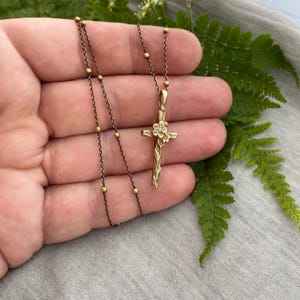 May include: A gold-toned cross pendant necklace. The cross has a floral design at the centre and is suspended from a delicate chain with small, round beads. The necklace is shown against a backdrop of green fern leaves and grey fabric.