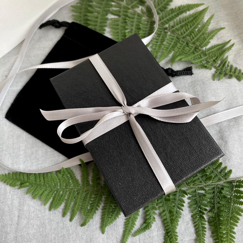 May include: A black gift box tied with a silver ribbon, set against green fern leaves. A black velvet pouch and a white ribbon add to the composition. The image presents a contrast of textures and colours, creating an elegant and natural aesthetic.