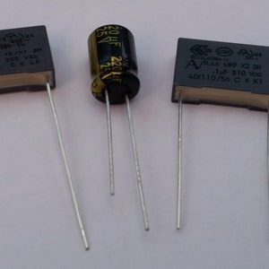 May include: Three black capacitors with silver leads. The capacitors are labeled with their capacitance and voltage ratings. The first capacitor is labeled "A/R.41 HEP YE/X1 SH 0.1 uF 300 V8C 40/110/68 CELE". The second capacitor is labeled "220uF 25V". The third capacitor is labeled "A/R46 MP X2 SH 1.0 uF 310 VoC 40/110/56 CKKI".