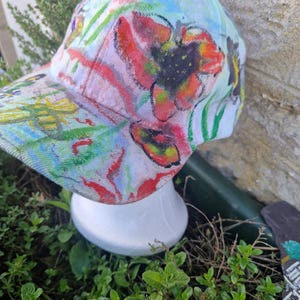 May include: A baseball cap with a colourful floral design. The cap has a light blue background with large red, yellow, and black flowers. Green leaves and stems are also visible. The cap is displayed on a white mannequin head.