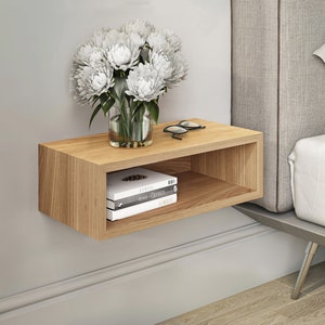 Peut inclure: Une étagère flottante en bois clair avec une petite étagère ouverte en dessous. L'étagère est fixée à un mur blanc et comporte un vase de fleurs blanches, une paire de lunettes et deux livres.