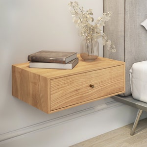 Peut inclure: Une table de chevet flottante en bois clair avec un seul tiroir et une poignée en laiton. La table de chevet est fixée à un mur blanc et a des livres empilés dessus. La table de chevet est à côté d'un lit avec une couette blanche et une tête de lit grise.