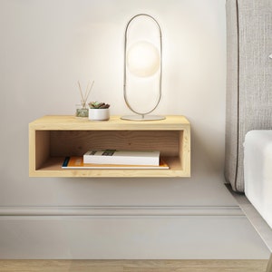 May include: A light wood floating shelf with a small potted succulent plant, a glass bottle with reeds, and a book on top. A silver lamp with a round white shade is on the shelf. The shelf is mounted on a white wall next to a bed with a gray headboard.