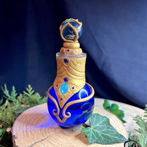 Op de afbeelding: Een decoratieve blauwe glazen fles met gouden accenten en een bolvormige stopper. De fles heeft een afgeronde basis en een gedetailleerd gouden ontwerp rond de hals. De stopper heeft een miniatuurlandschap in een blauwe bol.