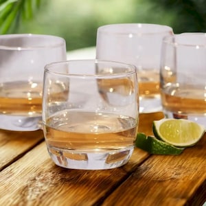May include: A set of four clear glass tumblers, each containing a light amber liquid. The glasses are arranged on a wooden surface, with a fresh lime wedge placed near the front glass. The design is simple, featuring a rounded base and rim.