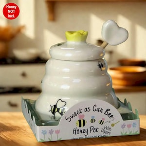 May include: A white ceramic honey pot with a yellow crown-shaped lid and a heart-shaped dipper. The pot is decorated with bee illustrations and the text "Sweet as Can Bee Honey Pot With Dipper". The product is displayed on a decorative tray.